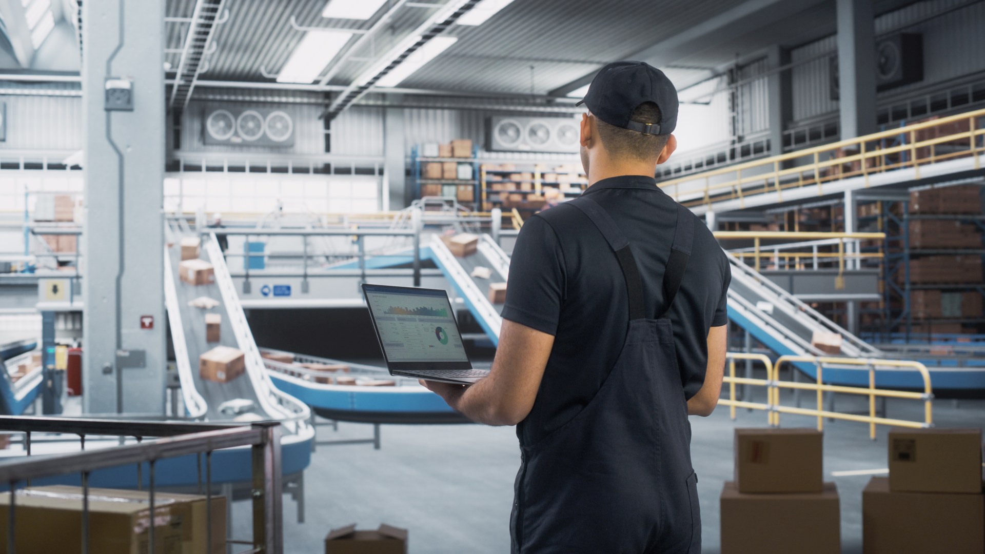Logistics Transportation Warehouse Facility: Associate Worker Using Laptop Computer in Distrubution Center, Monitoring the Conveyors Loading Online Ordered Product Boxes for Delivery to Customers.